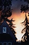 A darkened house and tall coniferous trees are silhouetted against a sky with orange smoke clouds — this house was heavily damaged in the Oakland Firestorm of 1991.