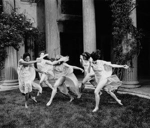 Black and white image of Isadora Duncan dancers at the Temple of Wings, Berkeley; five dancers are clustered, executing a running step sideways, with a hand in front of their faces, and back arm outstretched.