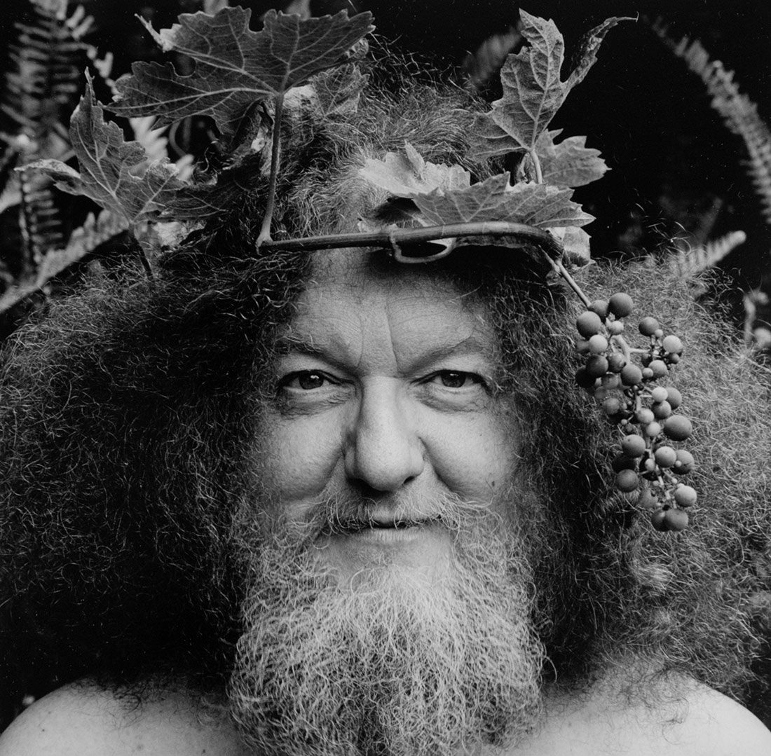 Black and white close-up of man's face with curly beard and hair that reach outward, his head crowned by a wreath of leaves and berries.