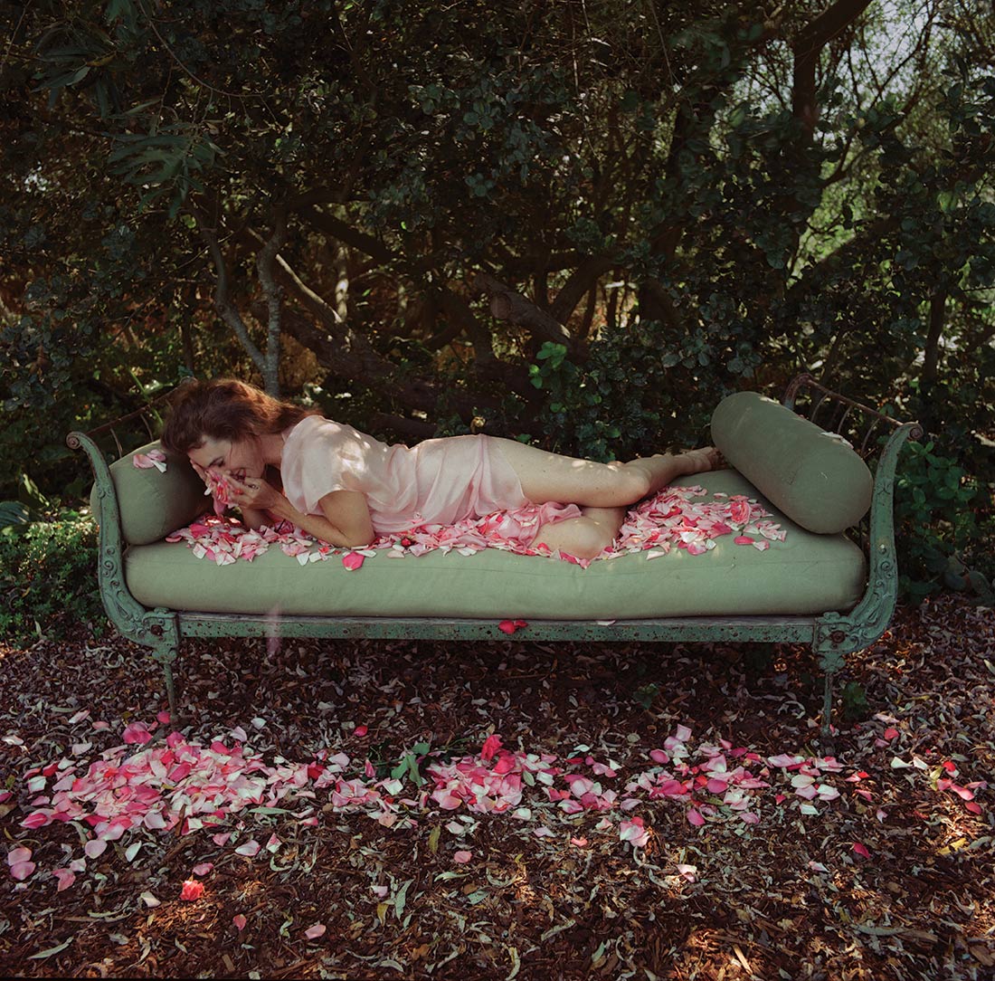 Color image of woman in silk garment reclining on an ornate bed in front of shrubbery; she is smiling into a handful of light and dark pink rose petals, and petals are strewn over her, the bed and the ground.
