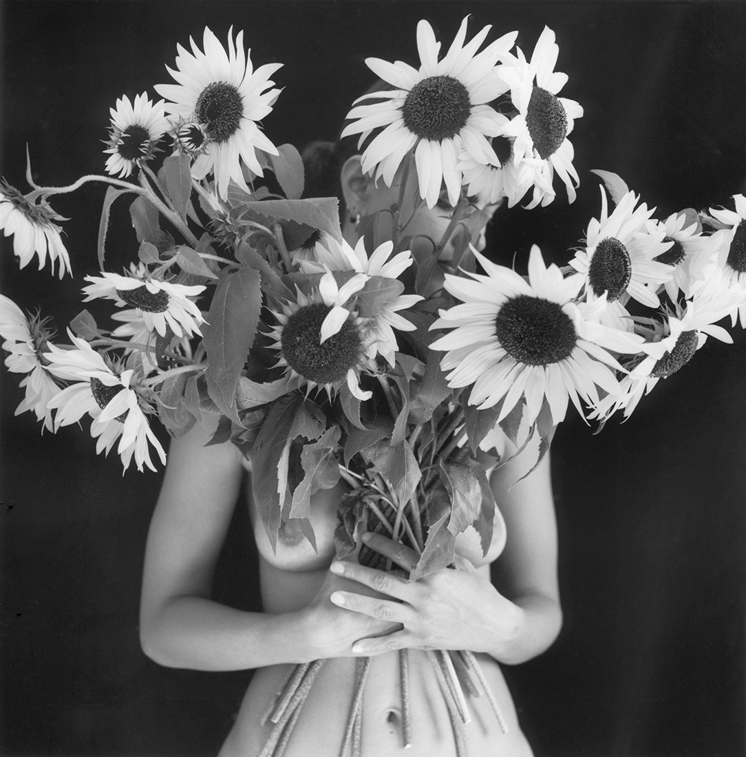 Black and white image of woman's holding bouquet of sunflowers on long stalks, the woman's face and breasts obscured, perceived as part of the bouquet.