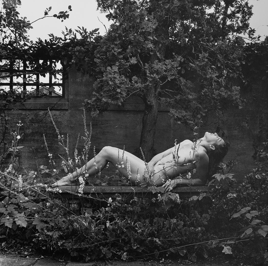 Black and white image of nude woman reclining on a garden bench, partially hidden by tall flower stalks; behind her is a small tree and a garden wall with a latticed window.