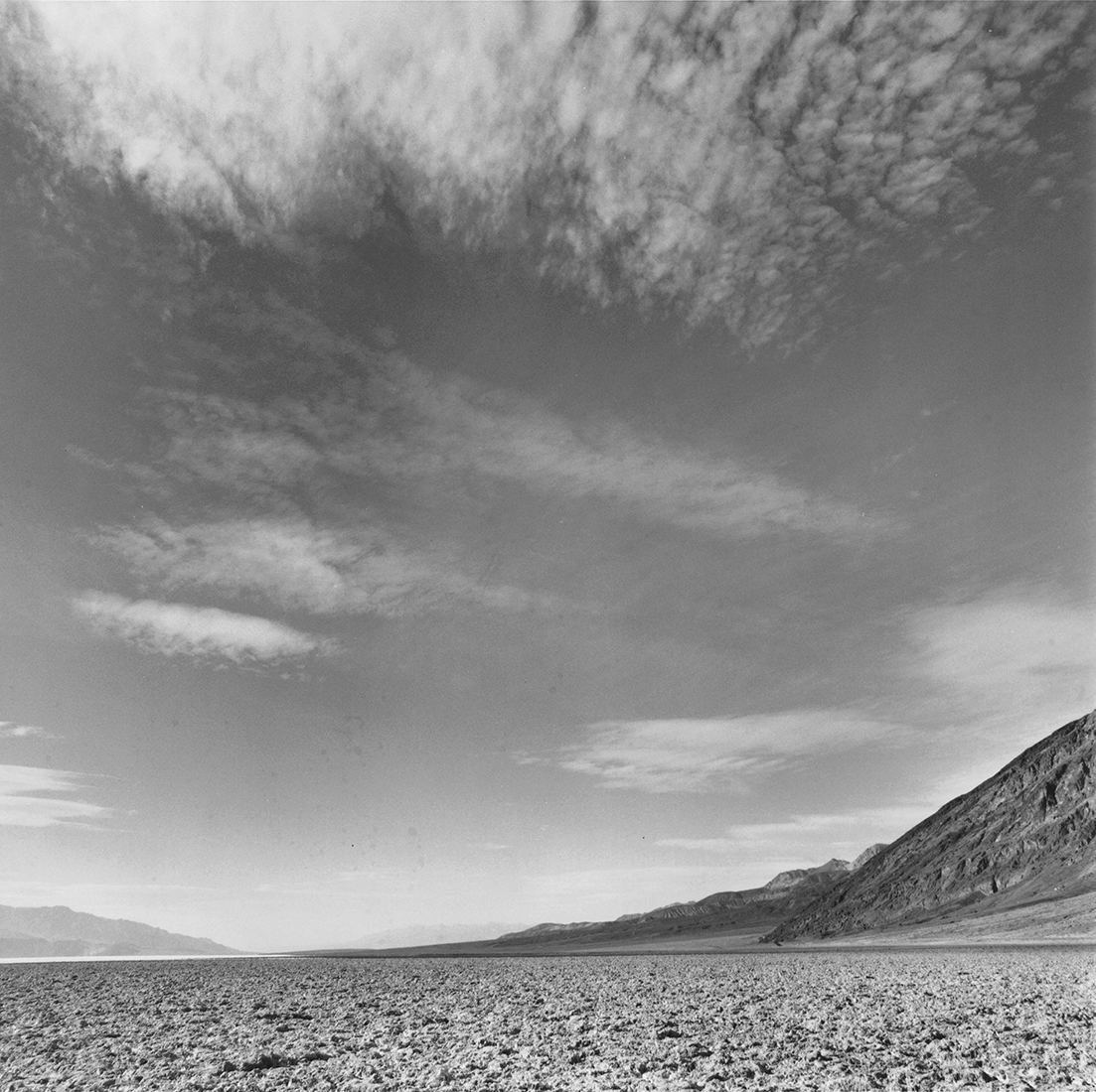 Cloud formations in a vast sky occupy most of the image with Death Valley salt flats and surrounding hills along the bottom