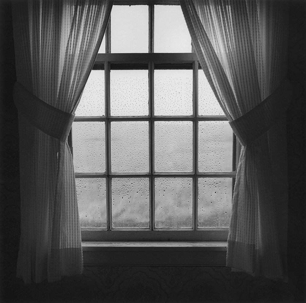Checked curtains are tied back to reveal raindrops on windowpanes, dark gray against the surrounding field below, luminous against the sky above.