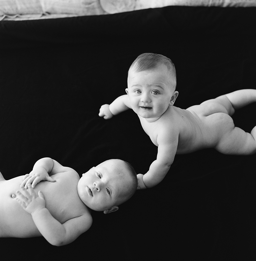 Black and white photograph of two human babies, light against a dark bedspread.