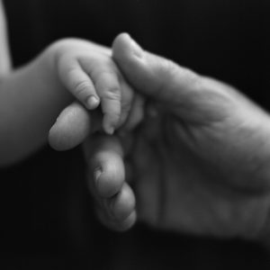 Black and white photograph of a large hand holding a tiny baby's hand between thumb and forefinger.
