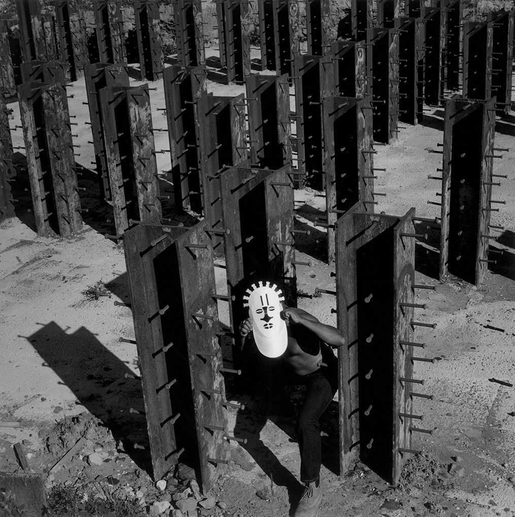 Black and white photograph of a man wearing a mask of the African god Ogun, seen among repeated steel beams protruding vertically from a concrete slab.